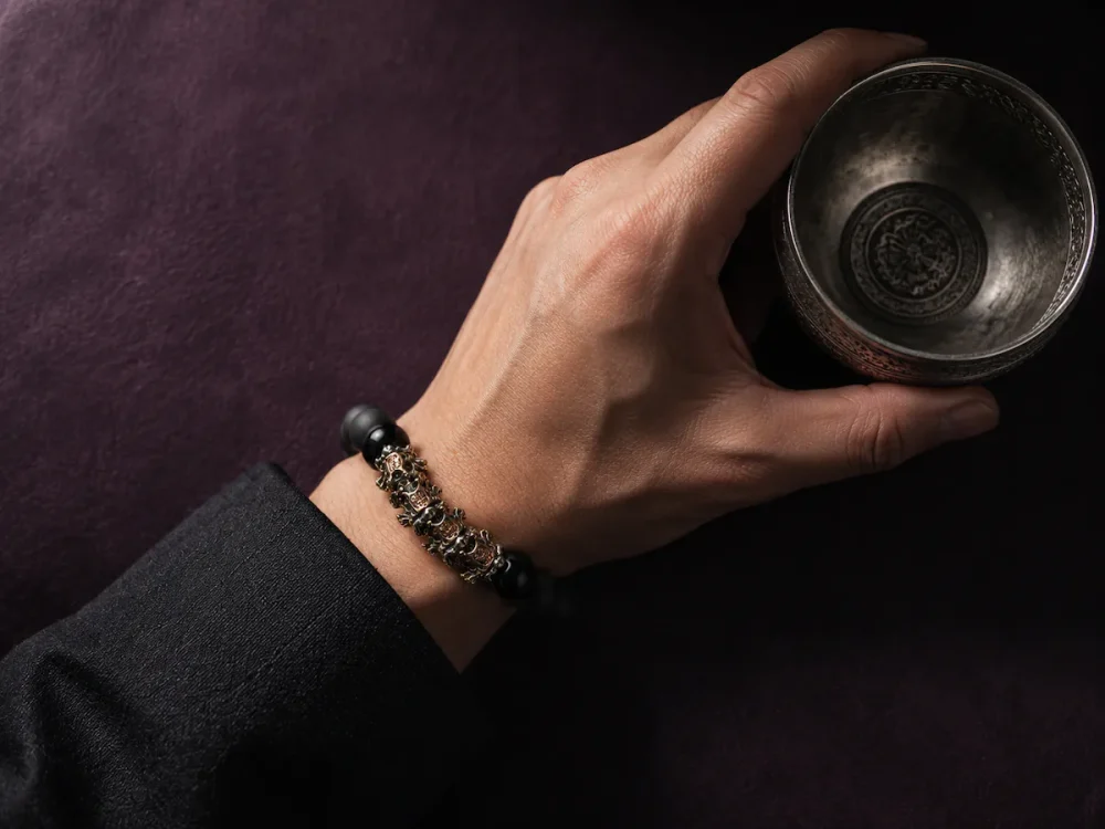 A man wearing the 9Purple dark zen obsidian bracelet with a charcoal-black tailored wool suit holding a vintage engraved silver tea bowl.