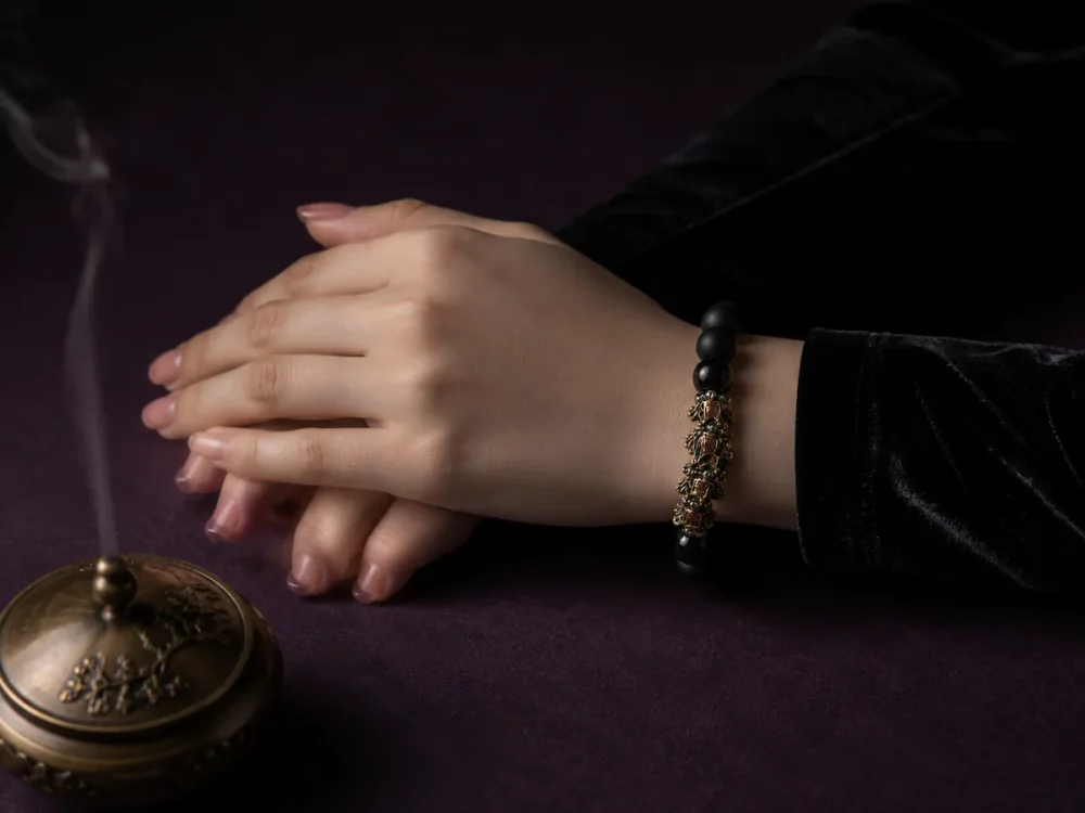 A woman elegantly wearing the 9Purple black obsidian bracelet with a black velvet sleeve, hands clasped next to a smoking vintage gold incense burner.