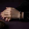 A woman elegantly wearing the 9Purple black obsidian bracelet with a black velvet sleeve, hands clasped next to a smoking vintage gold incense burner.