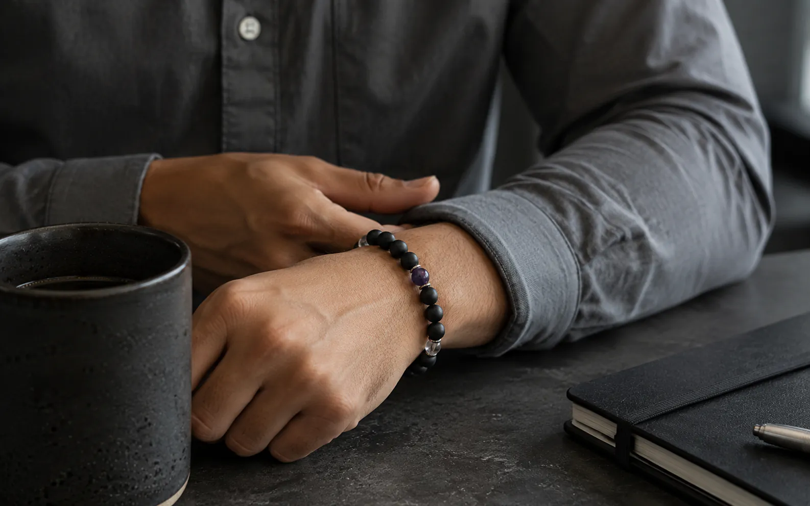 Minimalist luxury lifestyle shot showing a premium natural stone bracelet resting on an executive wrist over a clean gray workspace desk